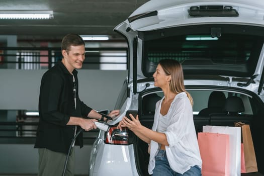 Young couple travel with EV electric car to shopping center parking lot charging in downtown city showing urban sustainability lifestyle by green clean rechargeable energy of electric vehicle innards