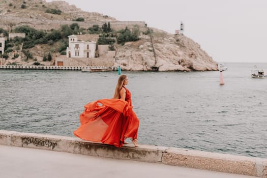 Side view a Young beautiful sensual woman in a red long dress posing on a volcanic rock high above the sea during sunset. Girl on the nature on overcast sky background. Fashion photo