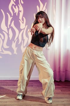A dancer performs a vibrant routine in a contemporary studio, wearing light-colored baggy pants and a fitted top. The backdrop features abstract flames, enhancing the energetic atmosphere.
