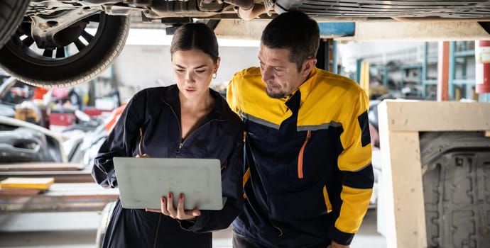 Two vehicle mechanic working together underneath lifted car, conduct car inspection with laptop. Automotive service technician in uniform carefully make diagnostic troubleshooting. Panorama Oxus