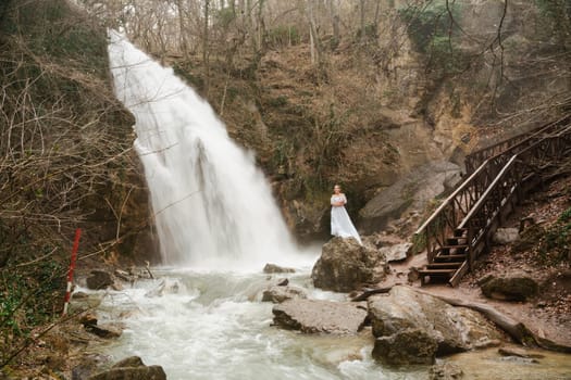 Happy woman in a white dress stands on a stone with a waterfall behind.