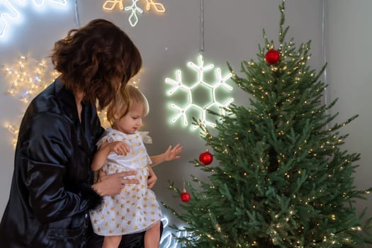 A mother with a 2-year-old daughter decorates the Christmas tree. Mom in a black suit, a girl in a white dress, her daughter hangs a red ball on the Christmas tree. Merry Christmas and New Year concept.