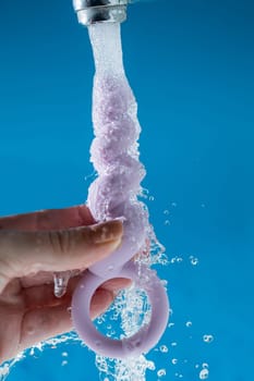 Woman holding lilac anal beads under running water on blue background. Sex toy hygiene concept