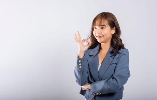 An infectious smile graces the face of an Asian girl in this cheerful studio portrait, as she confidently exhibits the 'OK' sign, symbolizing agreement and contentment against a white background.