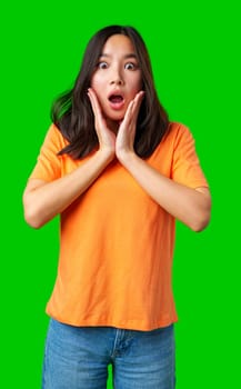 A young woman with long dark hair is wearing an orange shirt and denim jeans. She shows a surprised expression while holding her face, highlighting her astonishment.