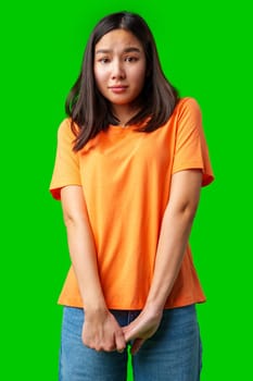 A young woman stands with a hesitant expression, wearing an orange shirt and denim jeans. The vibrant green background emphasizes her mixed emotions in a casual indoor atmosphere.