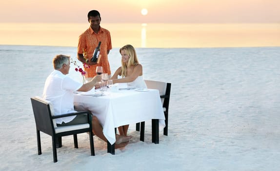 Couple, beach and dinner in sunset with waiter for romantic outdoor date, valentines day or anniversary. Man and woman with wine, cheers or celebration on table in relax or romance by ocean coast.