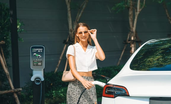 Young woman travel with EV electric car charging in green sustainable city outdoor garden in summer. Urban sustainability lifestyle by green clean rechargeable energy of electric BEV vehicle innards