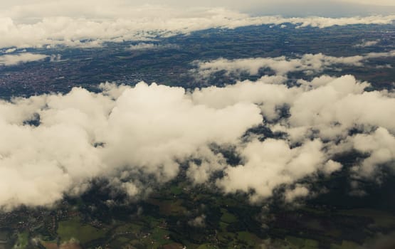 Beautiful aerial view of white cumulus clouds and the earth with fields, trees and settlements, close-up view from the top from an airplane window.