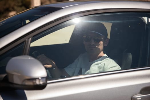 One young handsome Caucasian guy in a cap and sunglasses sits in a gray car behind the wheel on a sunny summer day, close-up side view.