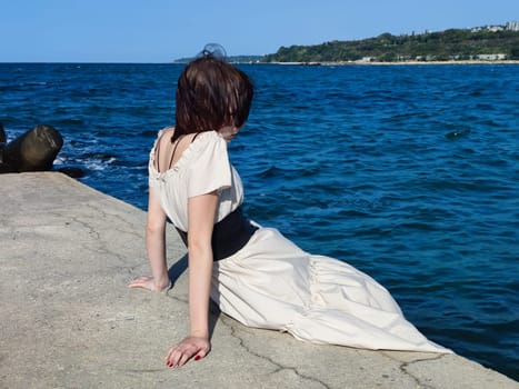 sad teenage girl in a white dress sits thoughtfully on the sea pier.