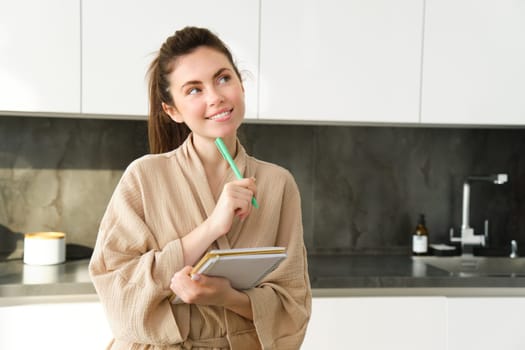 Portrait of beautiful modern woman, writing down grocery list, meal ideas in notebook, standing in the kitchen, wearing cosy bathrobe.
