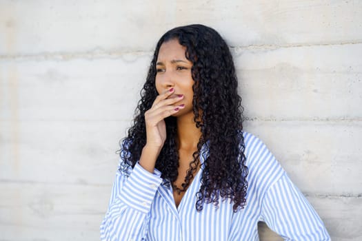 A thoughtful and contemplative young woman with curly hair stands gracefully against a raw concrete wall, appearing lost in deep thoughts, exuding a profound vibe of introspection and serene beauty