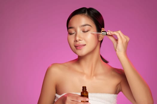 A woman with long hair applies skincare serum to her face while smiling. The vibrant pink background highlights her natural beauty during her daily skincare regimen.