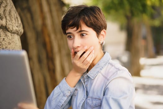 A young woman gasps in shock and disbelief as she gazes at her laptop, which she is using while sitting outdoors, capturing an exhilarating moment filled with surprise and joyful excitement