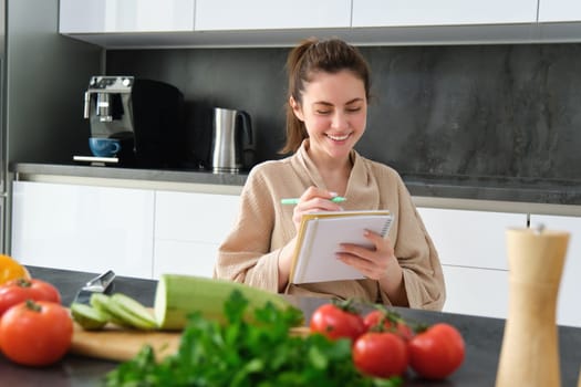 Portrait of beautiful, smiling young woman making list of meals, writing down recipe, sitting in the kitchen with vegetables, doing house errands.