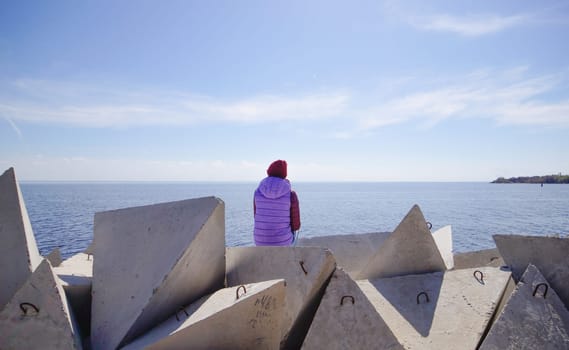 A young woman in a hat on a sunny spring day sits on the breakwaters and admires the sea. Girl in warm clothes on the breakwater