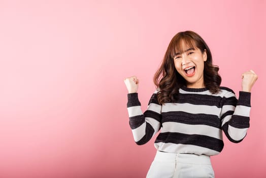 Portrait of a jubilant Asian woman with raised fists, celebrating her success with a happy expression. Studio shot isolated on a pink background, conveying victory and joy.