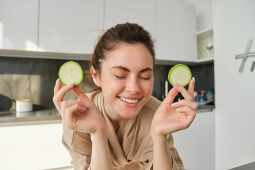 Portrait of happy, smiling young woman in the kitchen, cooking, chopping zucchini, holding vegetables and looking happy, preparing vegan food meal at home.