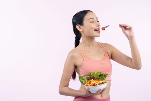 Young sporty Asian woman in sportswear holding salad bowl fill with vibrant of fruit and vegetable. Natural youthful and fit body lifestyle with balance nutrition on isolated background. Vigorous