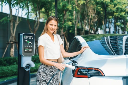 Young woman travel with EV electric car charging in green sustainable city outdoor garden in summer. Urban sustainability lifestyle by green clean rechargeable energy of electric BEV vehicle innards