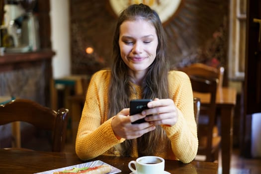 A young woman in a bright orange sweater smiles while using her smartphone in a warm caf setting with a coffee