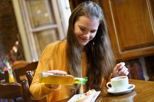 A cheerful young woman in a cozy caf drizzles olive oil over her dish while sipping coffee, highlighting a moment of culinary delight