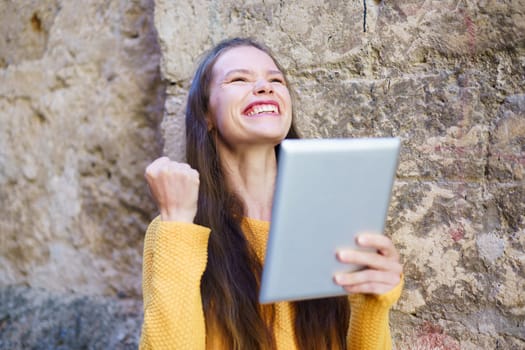 A joyful young woman celebrates her remarkable success while proudly holding up a tablet, expressing her excitement and happiness against the eyecatching backdrop of a rustic wall