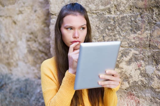 A young woman in a bright yellow sweater engages with her tablet while leaning against a rustic stone wall, blending contemporary tech with deep contemplation and personal reflection