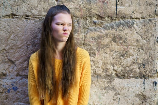 A young girl wearing a vibrant and brightly colored yellow sweater, displays a playful and humorous expression as she poses against a textured stone wall that adds depth to the scene