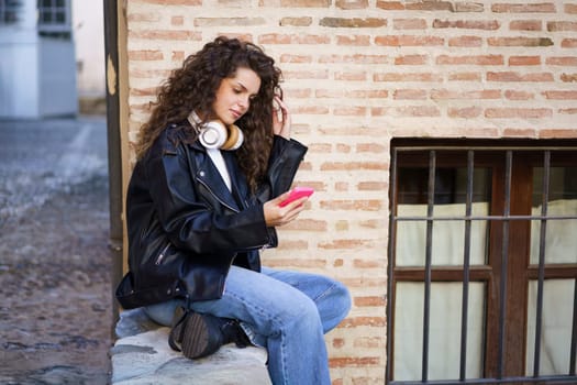 A fashionable young woman with curly hair sits on a stone wall, engrossed in her smartphone, reflecting modern youth culture in a vibrant urban setting filled with creativity and energy