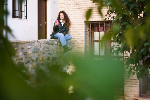 A carefree and lively young woman casually sits on a weathered stone wall, sipping a refreshing drink in a beautiful and scenic urban environment, surrounded by lush greenery all around her