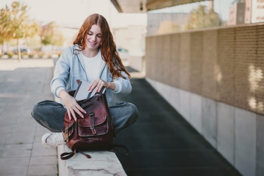 A young woman with long, flowing red hair is sitting comfortably on a stone ledge, leisurely opening her trendy brown backpack, all while enjoying the vibrant atmosphere of a modern urban setting