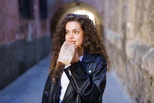 In a vibrant urban alley, a spirited young woman with lively curly hair, sporting a stylish leather jacket, indulges in a delicious snack, fully embracing a relaxed lifestyle admired by many others