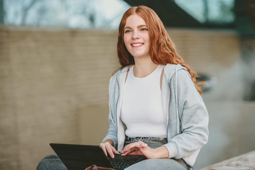 A young woman with long red hair sits outdoors with a smile, working on her laptop. She radiates positivity and productivity in a vibrant setting, reflecting her carefree yet focused lifestyle