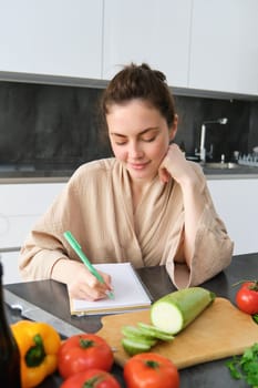 Vertical shot of smiling woman making notes, writing cooking recipe, creates groceries list, sits in the kitchen with vegetables and chopping board.
