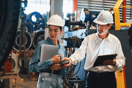 Factory engineer manager with assistant using laptop to conduct inspection of steel industrial machine, exemplifying leadership as machinery engineering inspection supervisor in metalwork manufacture.