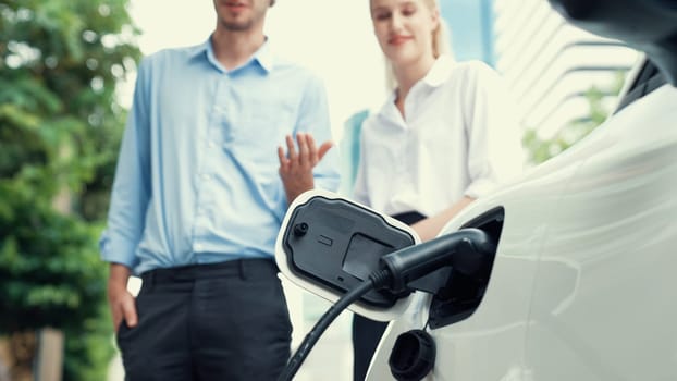 Progressive businessman and businesswoman install charger plug from charging station to electric car before driving around city center. Eco friendly rechargeable car powered by clean energy.