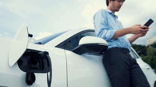 Suit-clad businessman with progressive ambition leaning on his electric vehicle while standing on a charging station with a power cable plug and a renewable energy-powered electric vehicle.