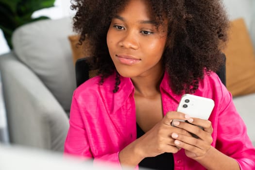 Young African businesswoman focusing information on smartphone and researching information data center on website pc and smartphone. Theme of place work in comfy room with plain color. Tastemaker.