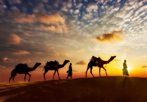 Travel background - two cameleers (camel drivers) with camels silhouettes in dunes of desert on sunset