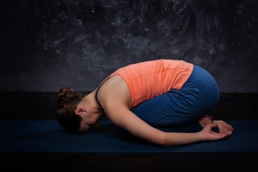 Beautiful sporty fit yogini woman practices yoga asana balasana (child's pose) - resting pose or counter asana for many asanas on dark background