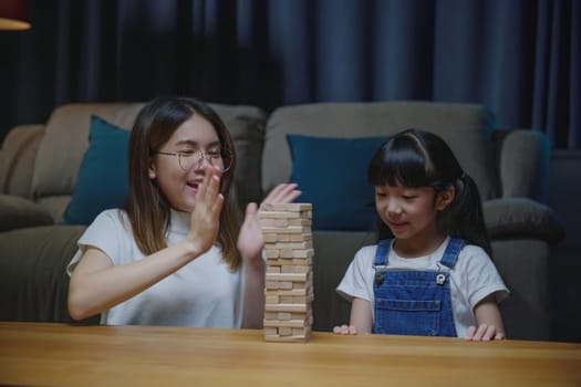 Asian young mother playing game in wood block with her little daughter in home living room at night time, Smiling woman help teach child play build constructor of wooden blocks, education