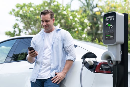 Modern eco-friendly man recharging electric vehicle from EV charging station, using Innovative EV technology utilization for tracking energy usage to optimize battery charging on smartphone.Synchronos