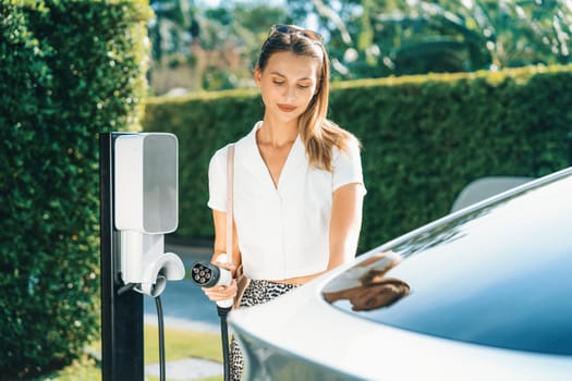 Young woman travel with EV electric car charging in green sustainable city outdoor garden in summer. Urban sustainability lifestyle by green clean rechargeable energy of electric BEV vehicle innards