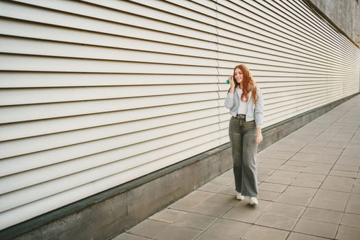 A stylish woman walks a modern sidewalk, focused on her phone, surrounded by sleek architecture. She represents leisure and individuality in trendy jeans and sneakers, enjoying the vibrant city