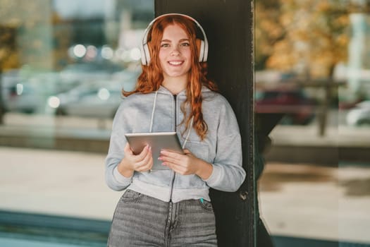 A cheerful young woman featuring curly red hair and trendy headphones beams radiantly with happiness as she holds a sleek tablet, thoroughly immersed in her music in a lively, vibrant outdoor setting