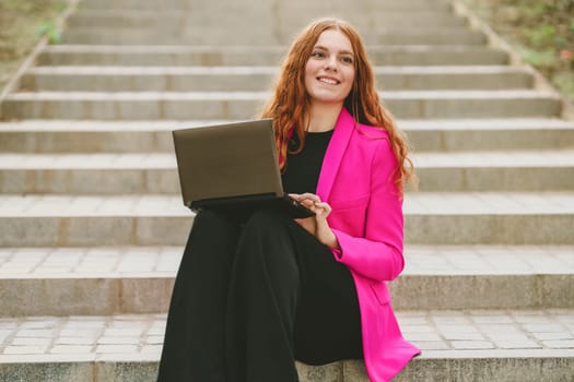 A stylish young woman in a vibrant pink blazer sits on colorful steps, focused on her laptop, blending professionalism with creativity, inspiring others with her dedication and modern lifestyle
