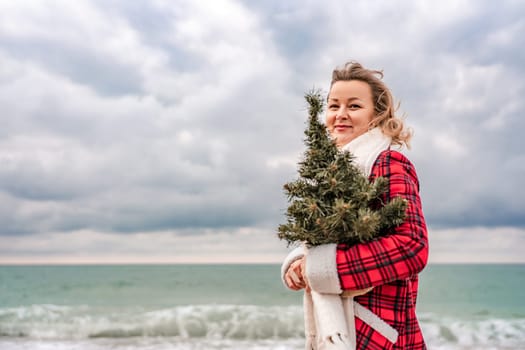 Blond woman holding Christmas tree by the sea. Christmas portrait of a happy woman walking along the beach and holding a Christmas tree in her hands. Dressed in a red coat, white dress