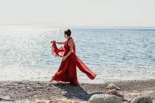 Side view a Young beautiful sensual woman in a red long dress posing on a rock high above the sea during sunrise. Girl on the nature on blue sky background. Fashion photo.